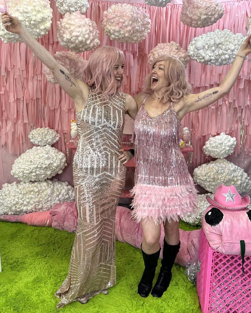 Elissa and Melissa in sequins and pink wigs, arms up, in a pink photo booth