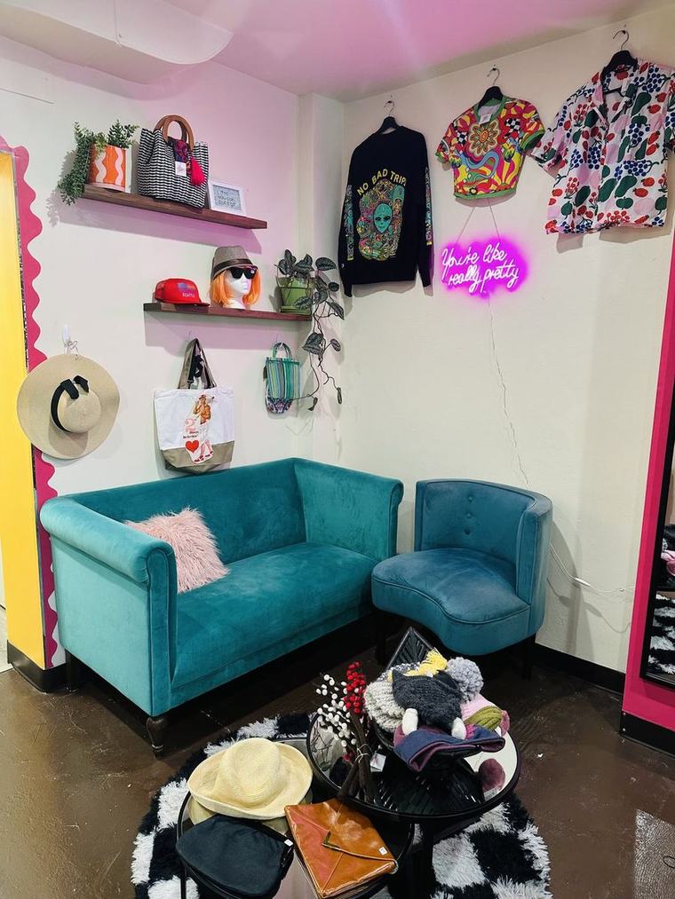 The teal velvet loveseat and chair by the racks, hats on the table, a pink neon sign on the wall